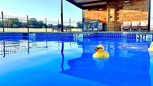 Une piscine avec un canard portant des lunettes de soleil, un bâtiment en bois avec une pancarte indiquant « PISCINE », et un espace clos avec des chaises et des tables.
