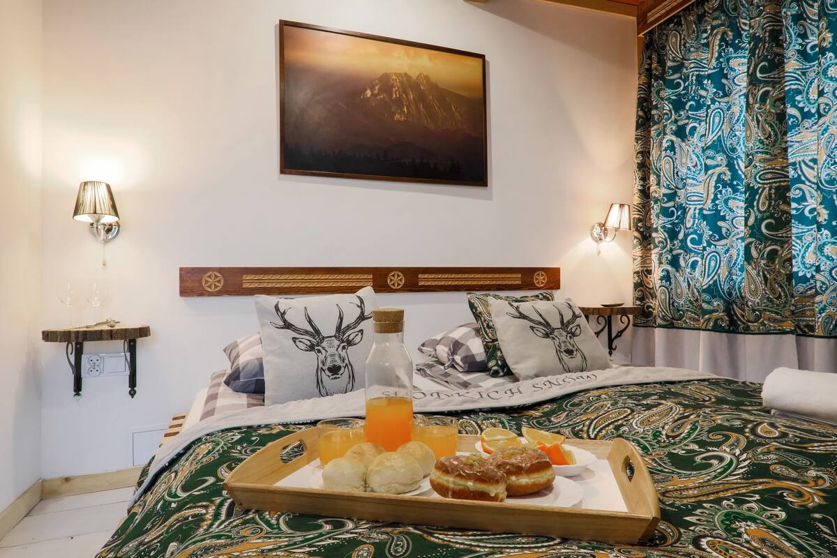 A neatly made bed with a green patterned bedspread, a tray of breakfast items including pastries and orange juice, and a mountain landscape painting above the bed.