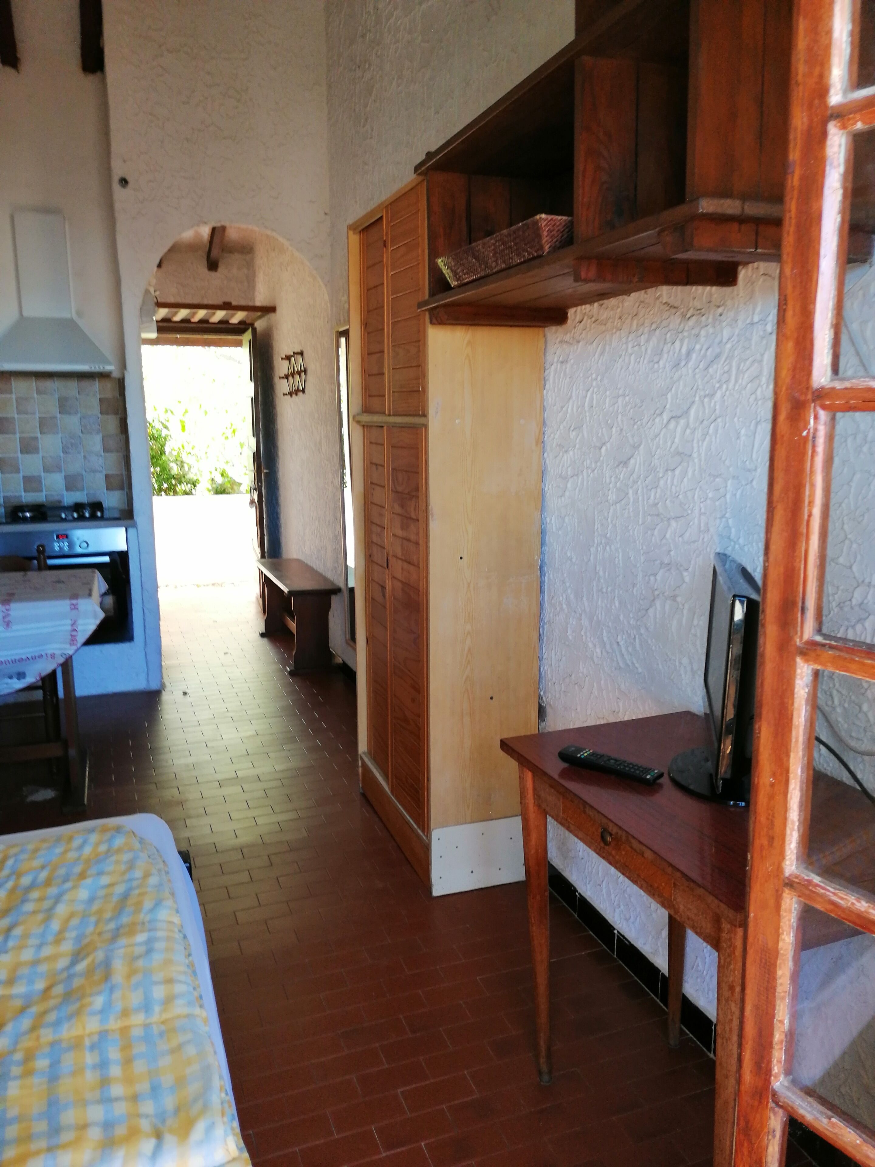 Cocina compacta con estufa y lavabo, una mesa de comedor de madera con control remoto, y una cama con manta de cuadros.