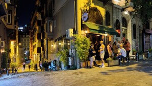 A nighttime street scene with people walking and sitting outside a building with a sign that reads "ELLEK."