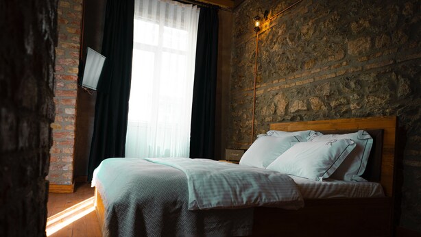 A bedroom with a wooden bed, a brick wall, a window with curtains, and a wall-mounted light fixture.