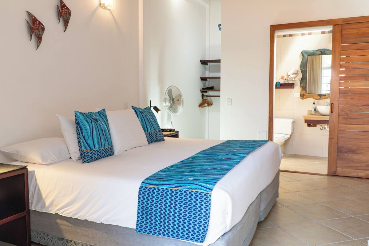 Ein Schlafzimmer mit Bett, Nachttisch, Lampe und Blick ins Badezimmer.