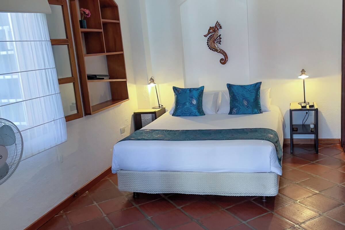 Ein Schlafzimmer mit einem Bett, zwei Nachttischen mit Lampen, einem Holzregal, einer Wanddekoration mit einem Seepferdchen und einem Fenster mit Vorhängen.