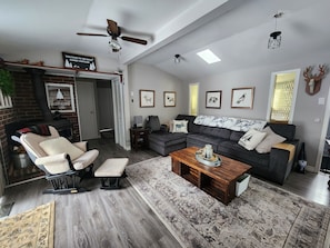 A spacious living room with a fireplace, a ceiling fan, and a large window.