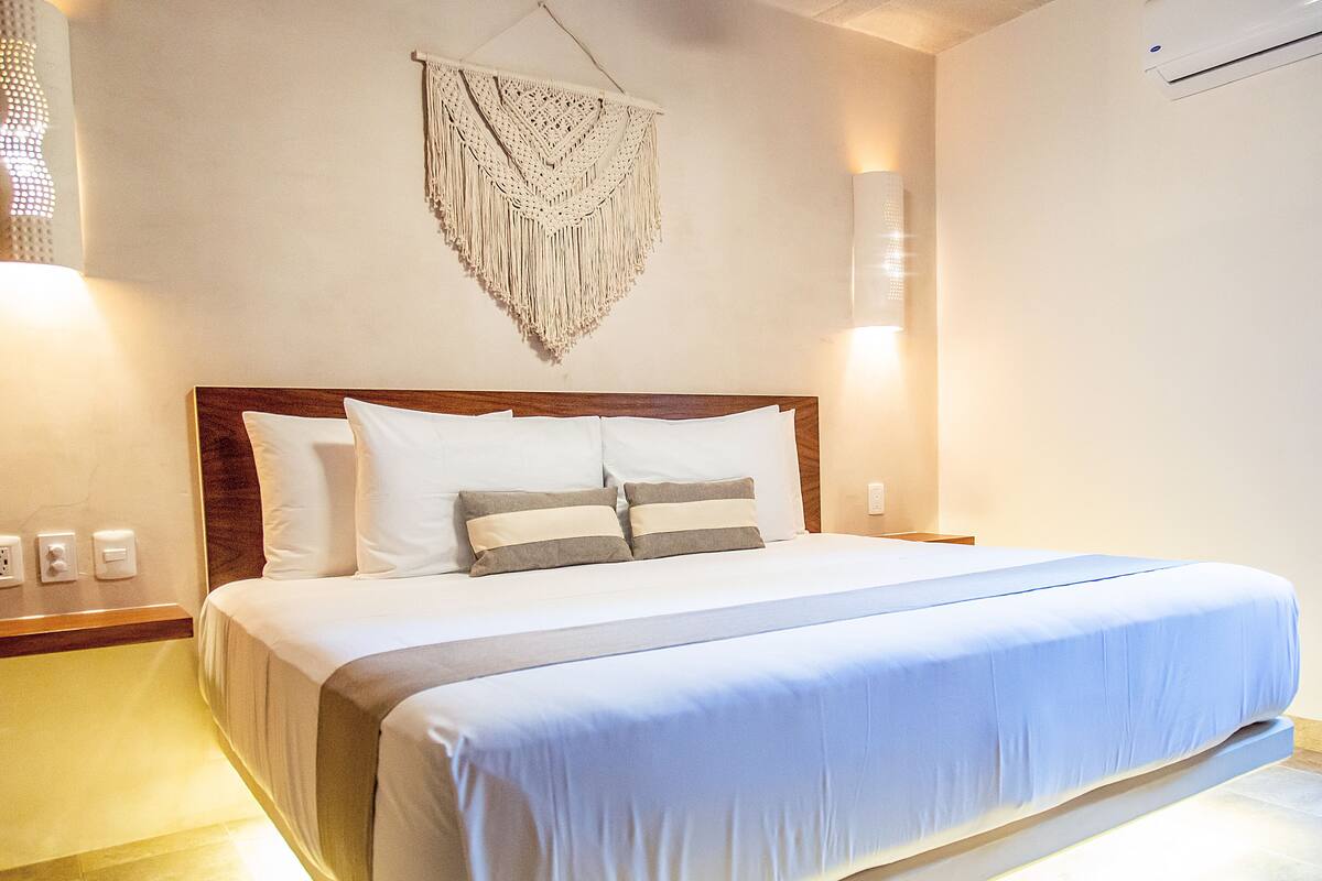 A neatly made bed with white and gray linens, a wooden headboard, and a macrame wall hanging.