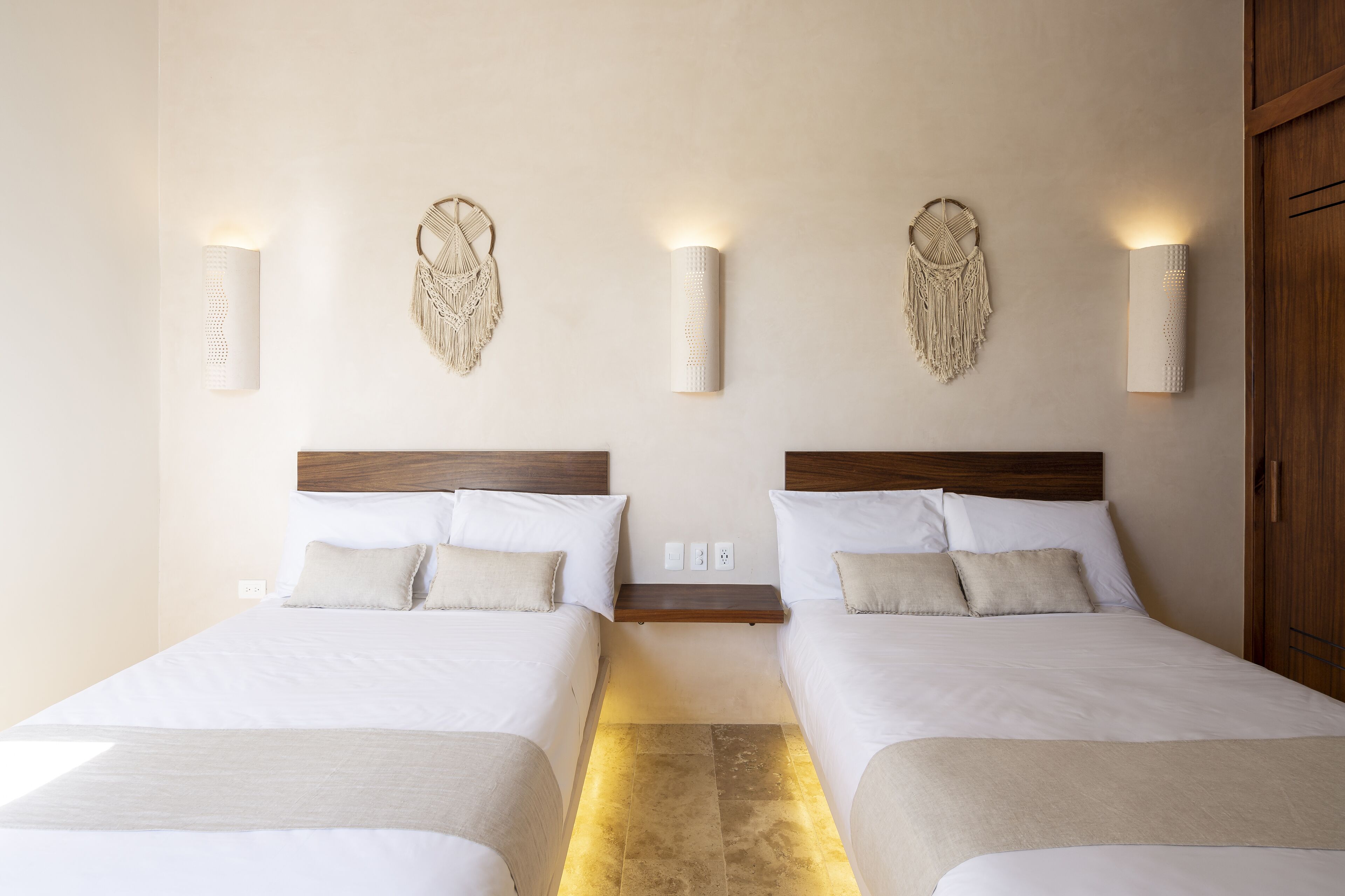 A hotel room with two beds, a wooden headboard, wall-mounted macrame wall art, and a small wooden shelf between the beds.