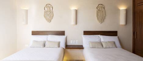A hotel room with two beds, a wooden headboard, wall-mounted macrame wall art, and a small wooden shelf between the beds.