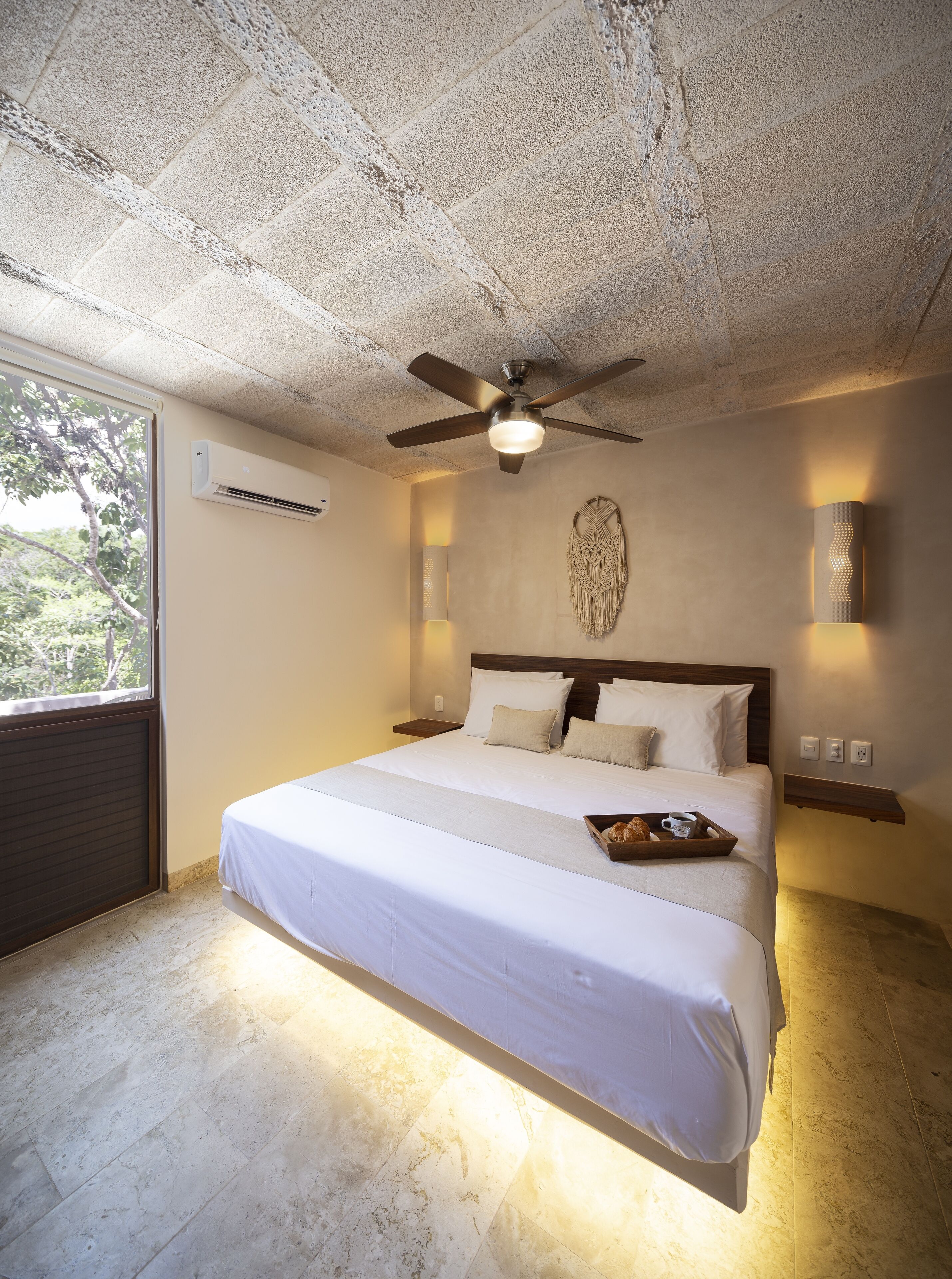 A bedroom with a large bed, a ceiling fan, an air conditioning unit, a wall-mounted light, and a window with a view of trees.