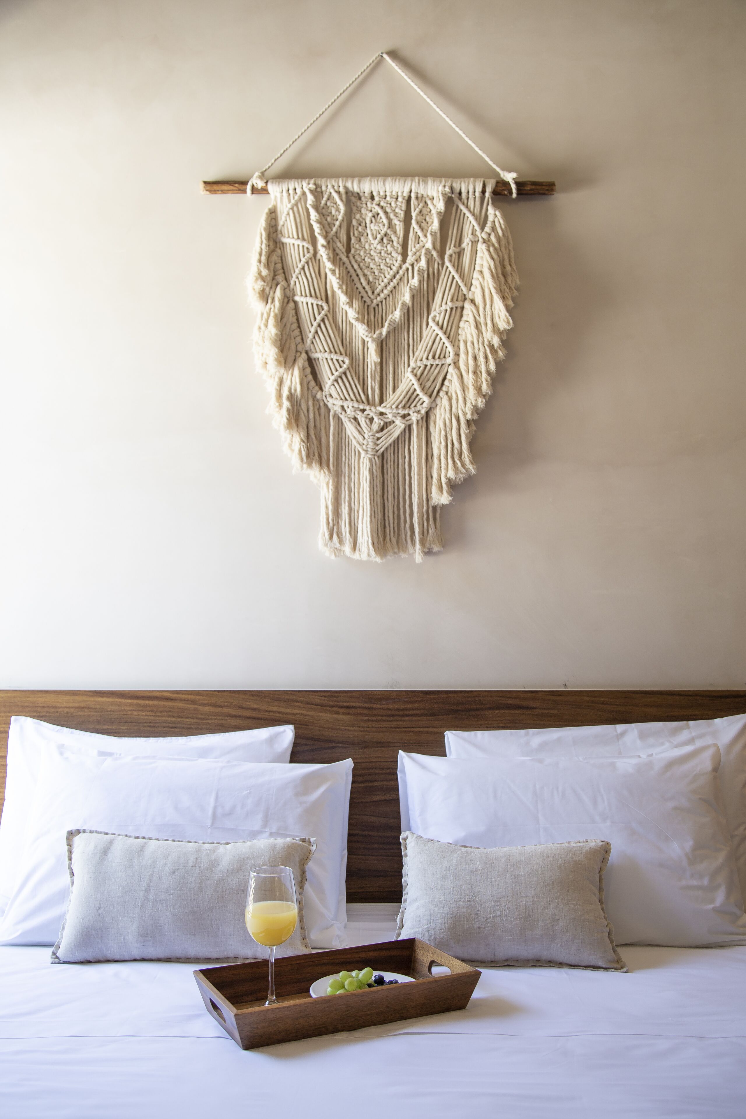 Een bed met witte beddengoed, een macramé wanddecoratie, en een dienblad met een glas sap en fruit.