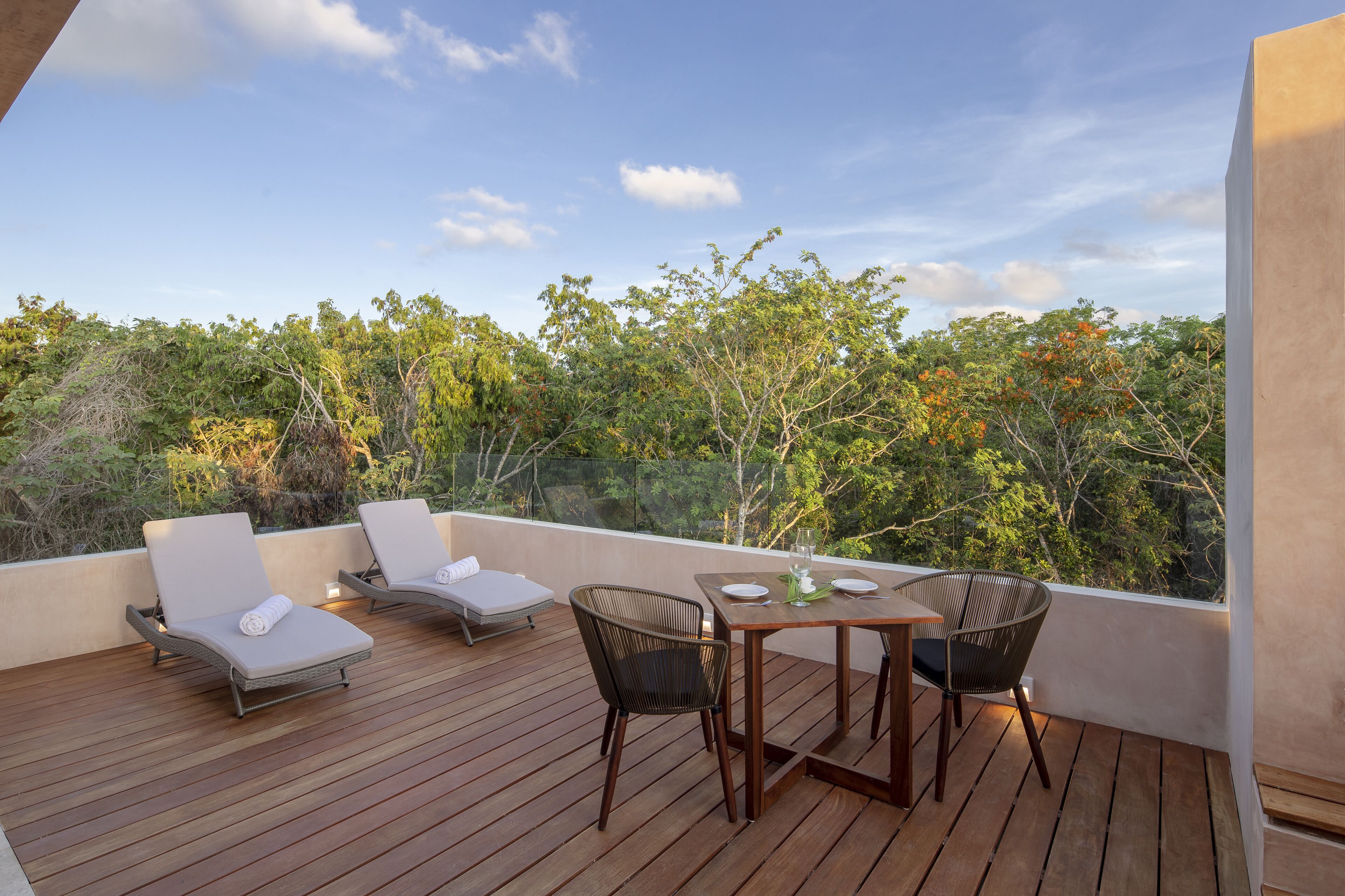 A wooden deck with two lounge chairs and a table, overlooking a forested area.