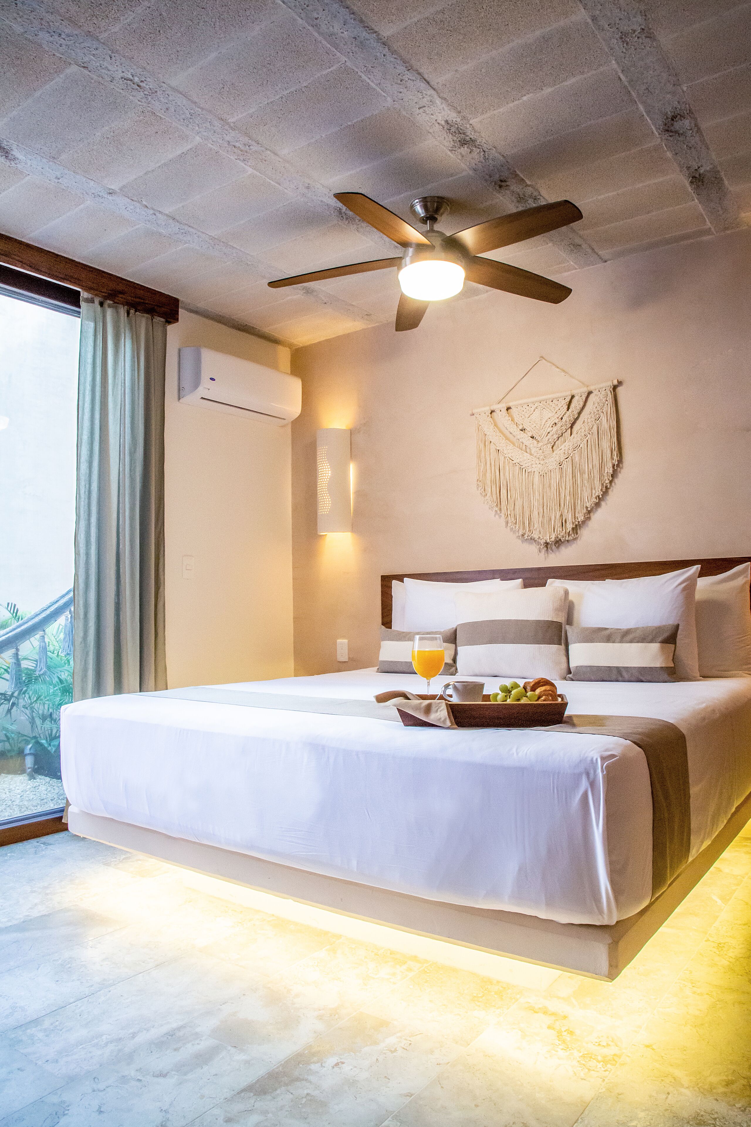 A hotel room with a large bed, a ceiling fan, a wall-mounted air conditioner, a macrame wall hanging, and a window with curtains.