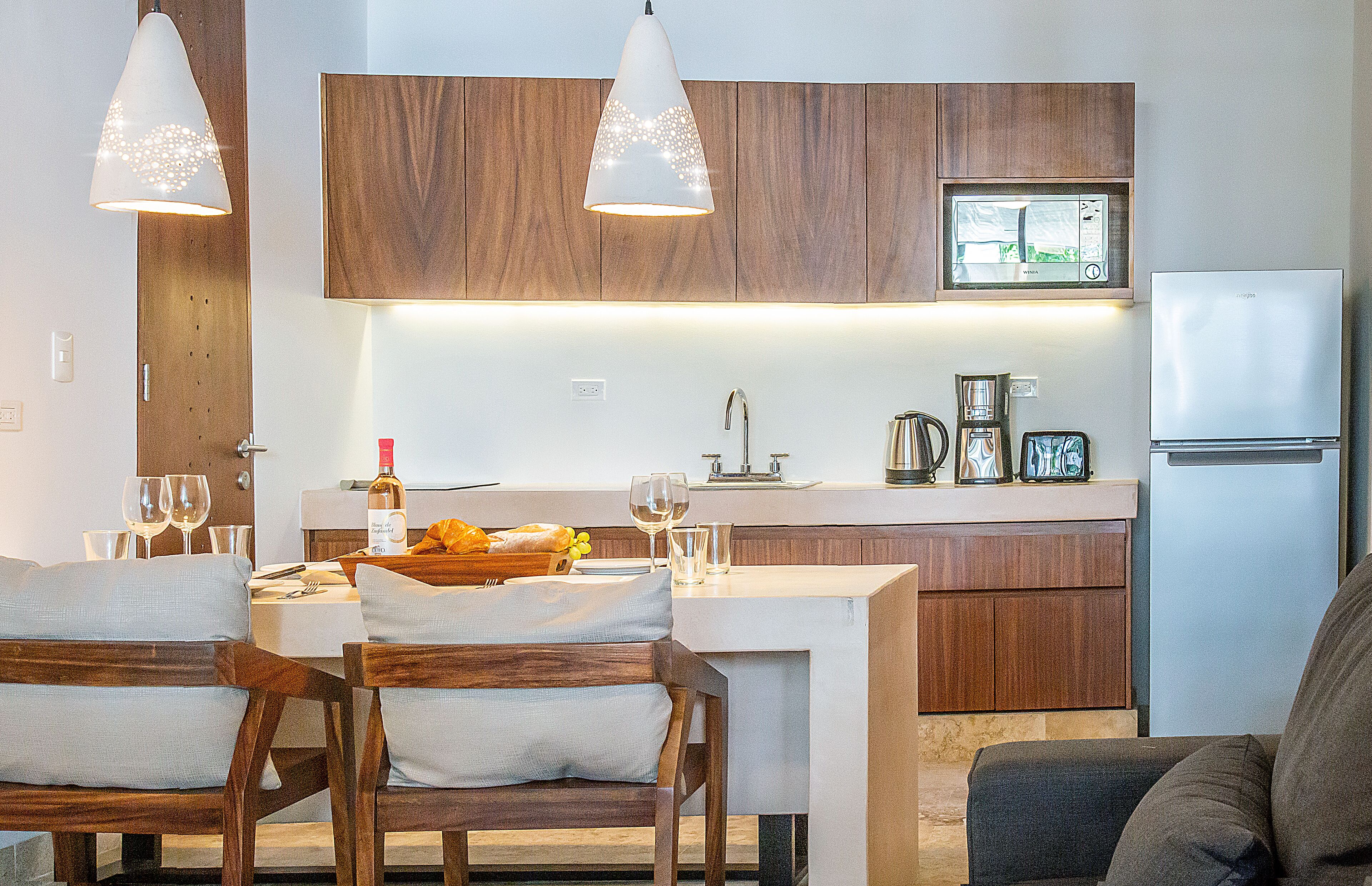 Eine moderne Küche mit Holzschränken, integriertem Mikrowellenherd und Kühlschrank. Es gibt einen Esstisch mit Stühlen sowie eine kleine Kücheninsel mit einer Weinflasche und Gläsern.