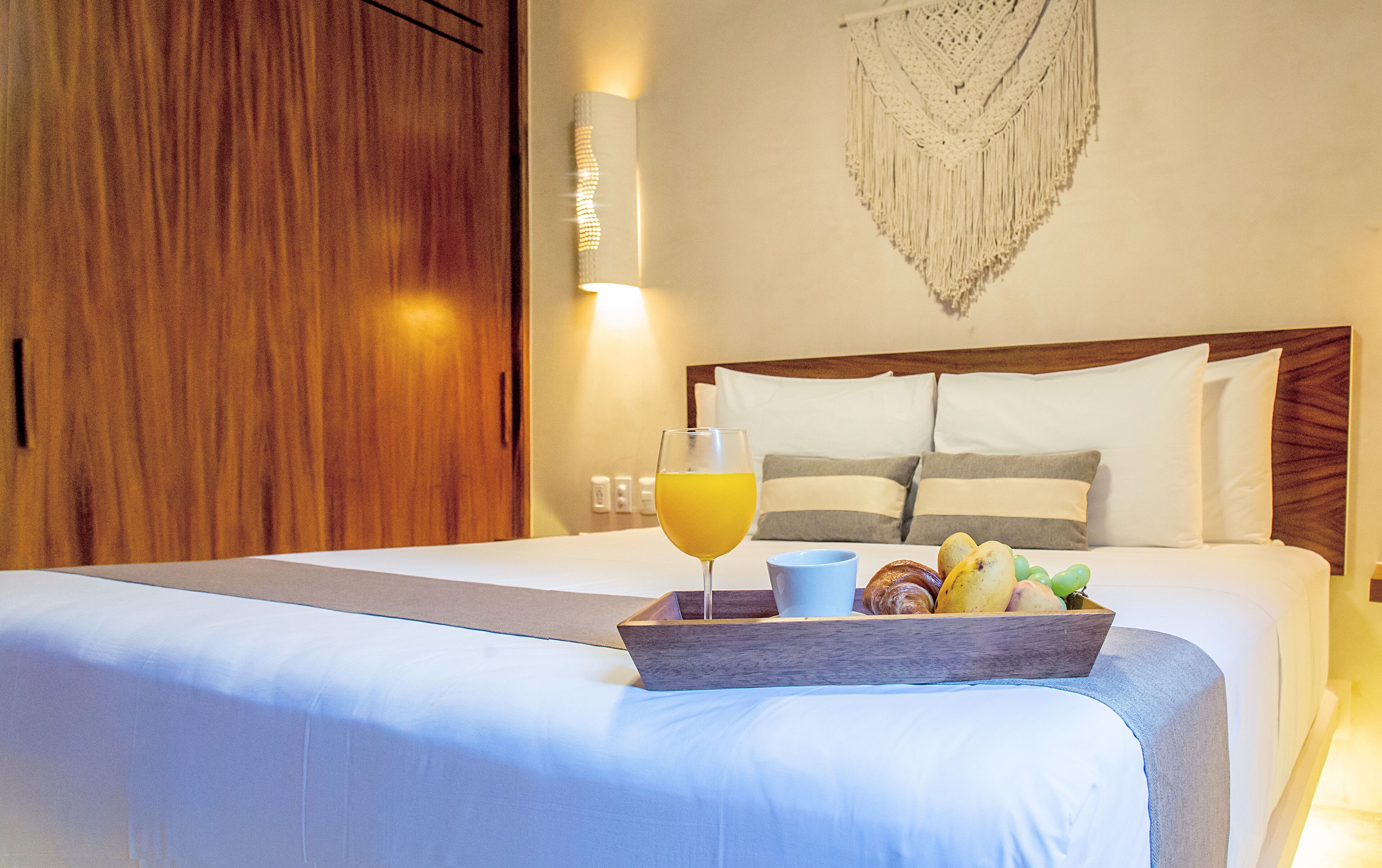 A hotel room with a neatly made bed, a tray of breakfast including croissants, fruits, and a glass of orange juice, and a wall-mounted light fixture.