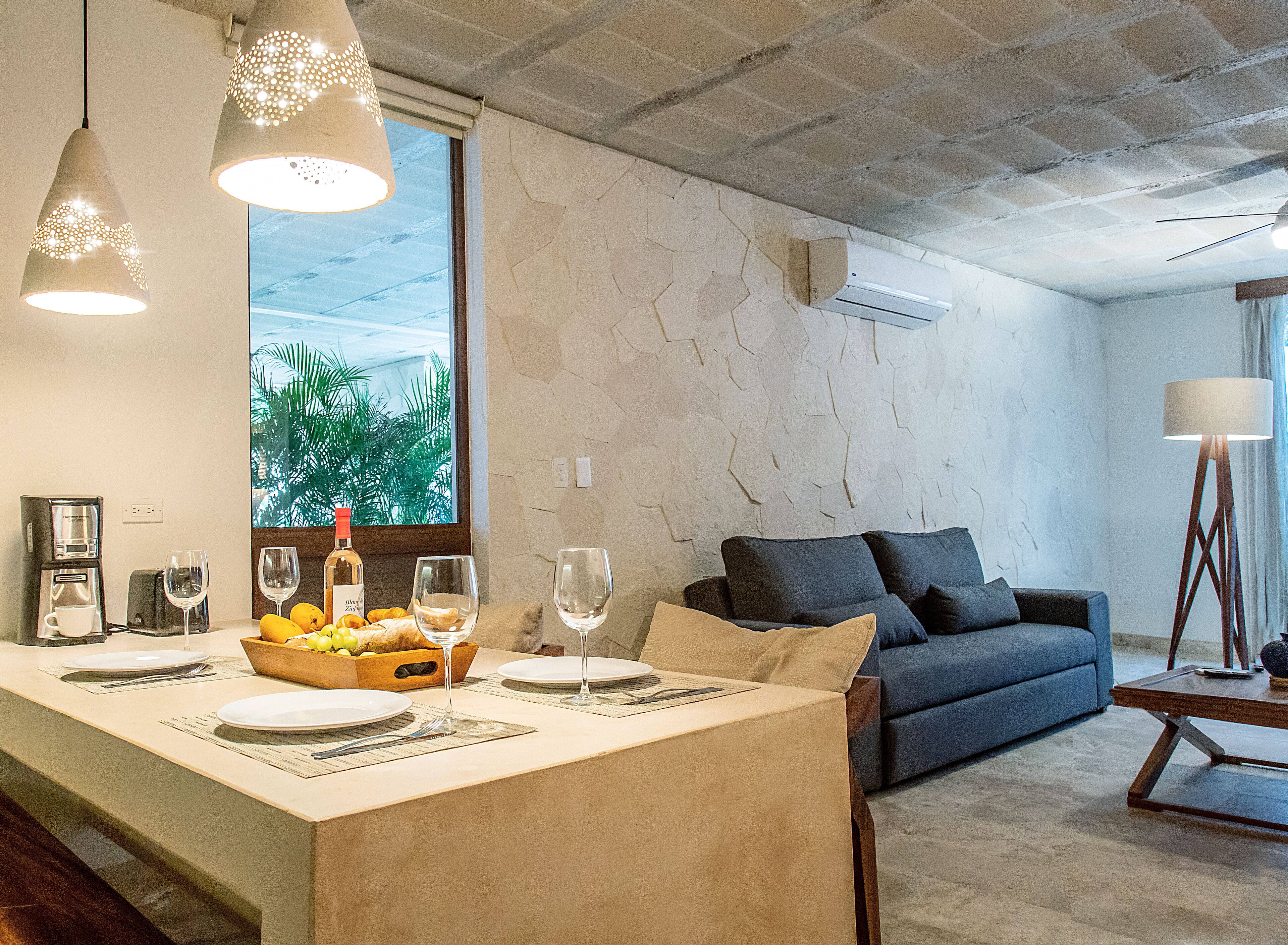 A modern living room with a sofa, a dining area with a table set for two, and a large window with a view of greenery.