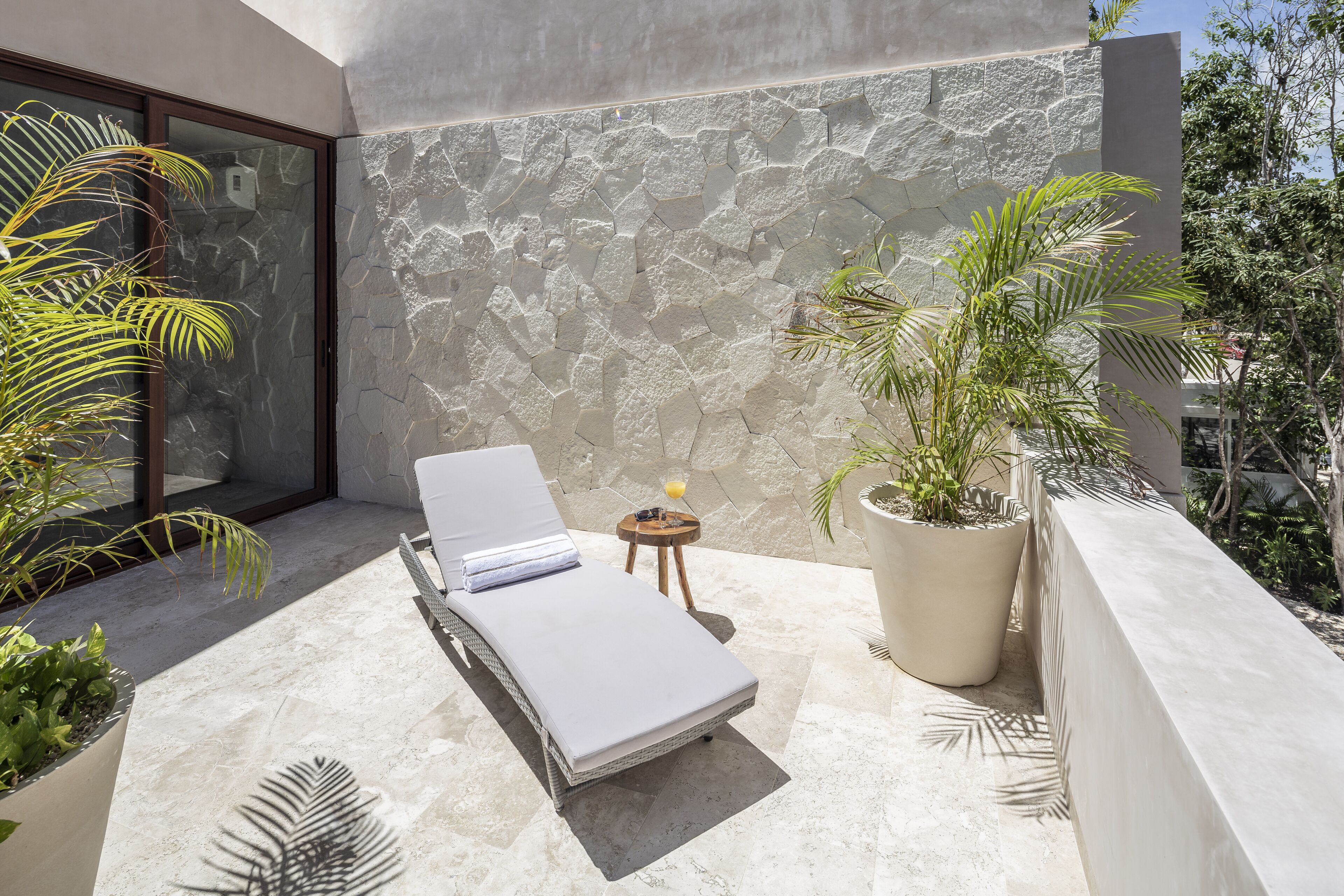 A sunlit balcony with a lounge chair, a potted plant, and a stone wall.