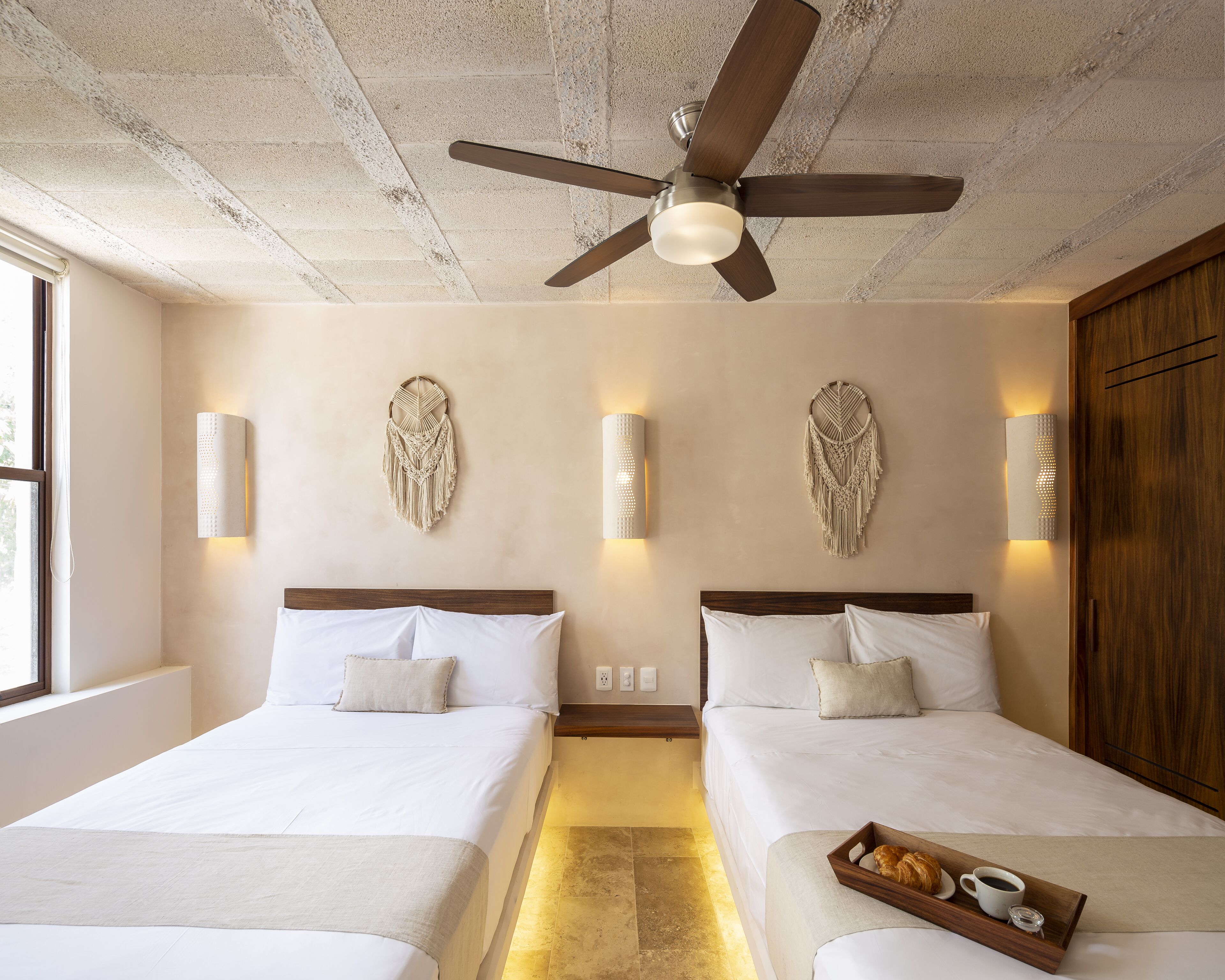 A hotel room with two beds, a ceiling fan, wall-mounted macramé wall art, and a tray with croissants and coffee on the bed.