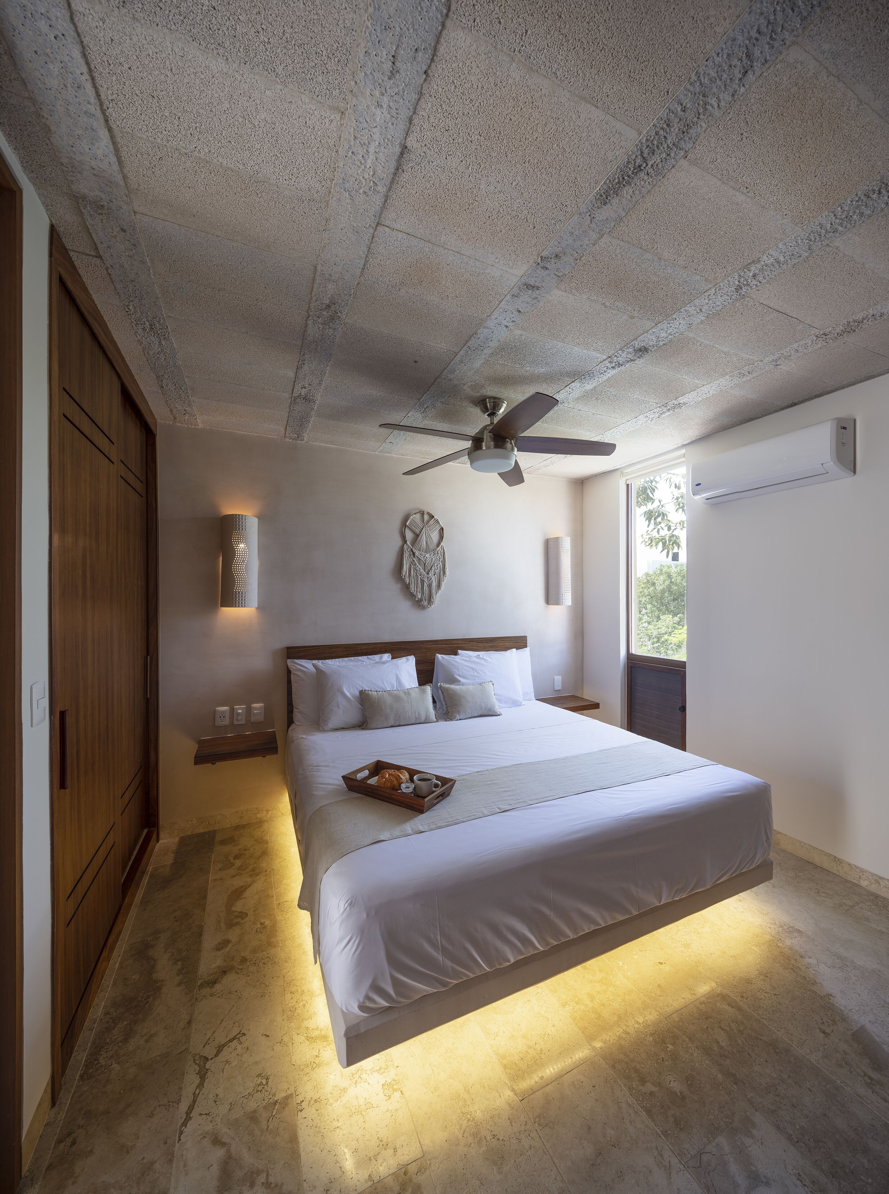 A bedroom with a large bed, a ceiling fan, a wall-mounted air conditioner, and a window with a view of greenery.