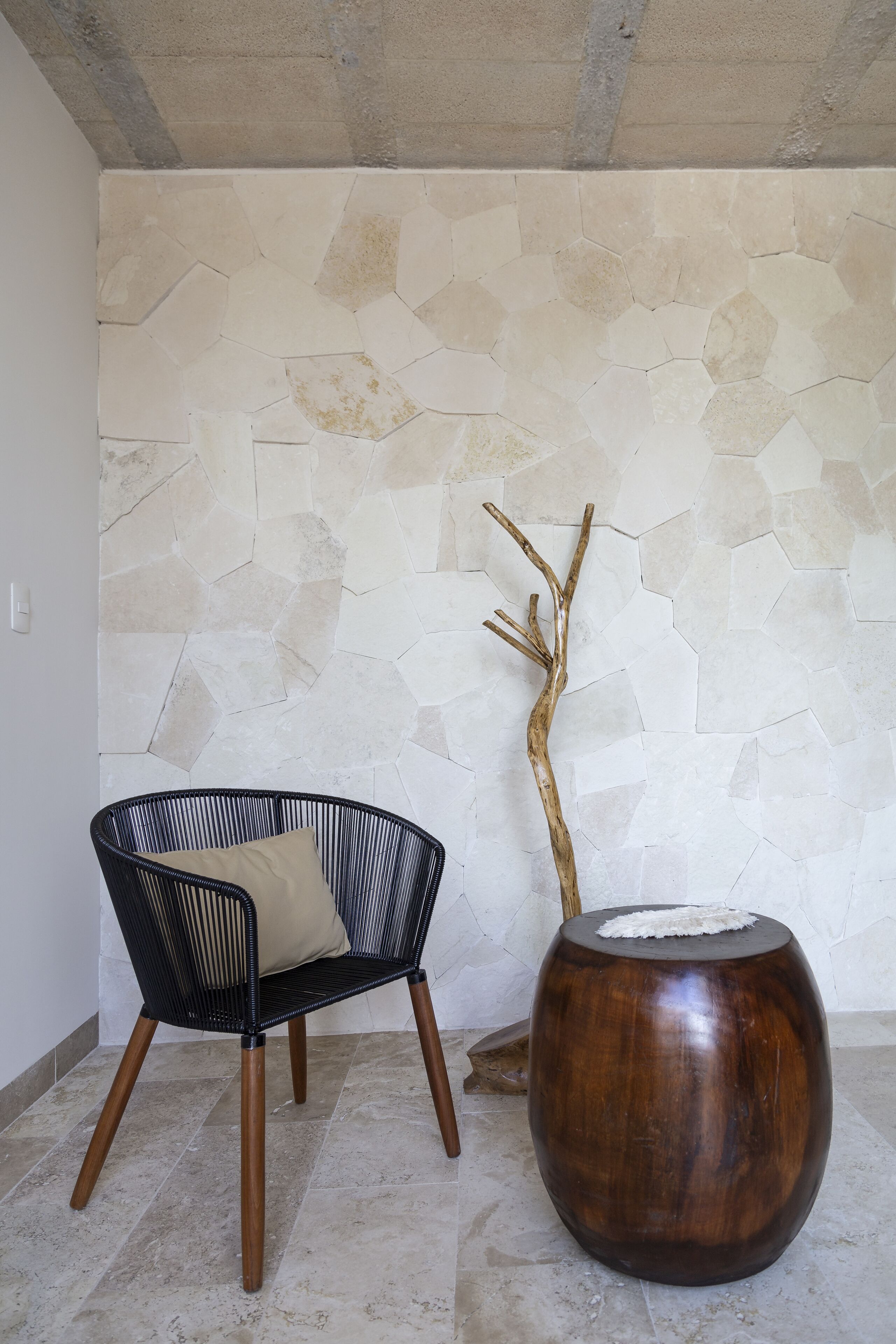 A room with a stone wall, a black woven chair with a cushion, and a wooden table with a branch decoration.