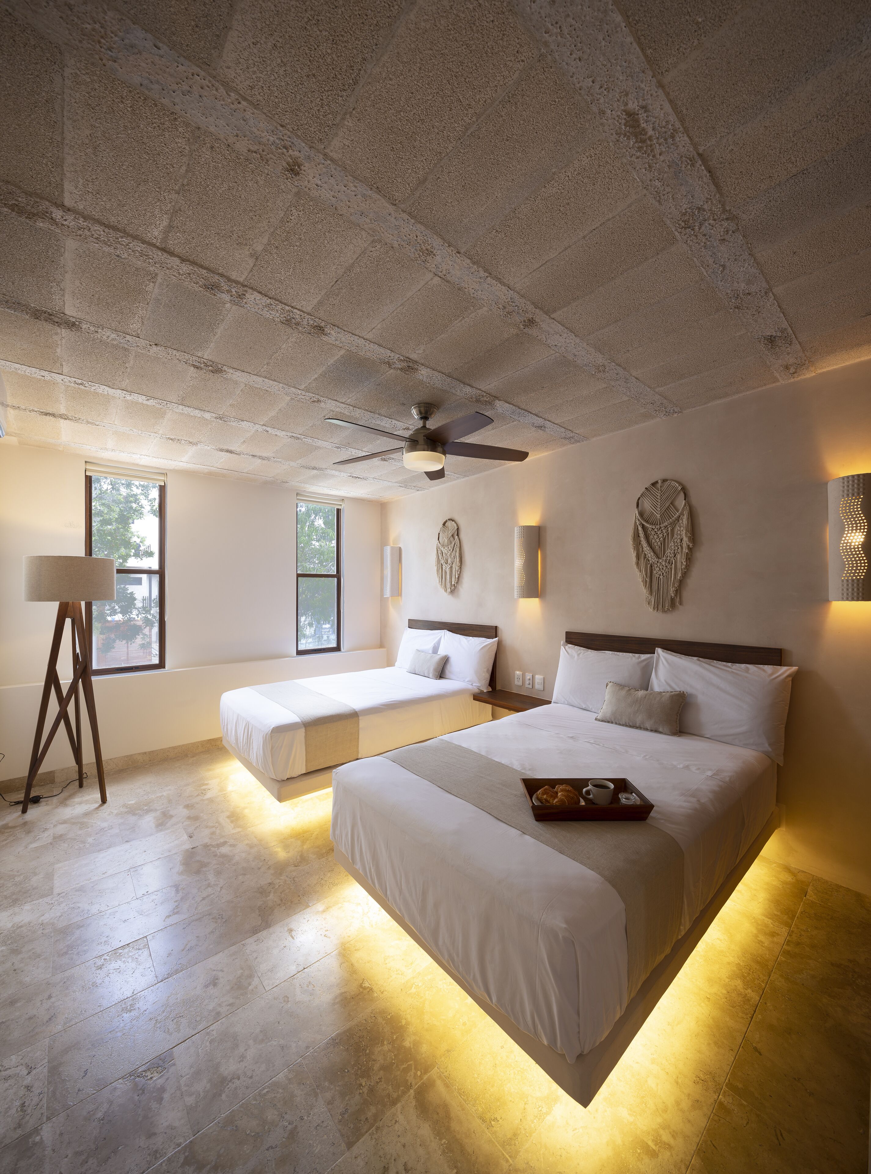 A hotel room with two beds, a ceiling fan, a standing lamp, and a large window.