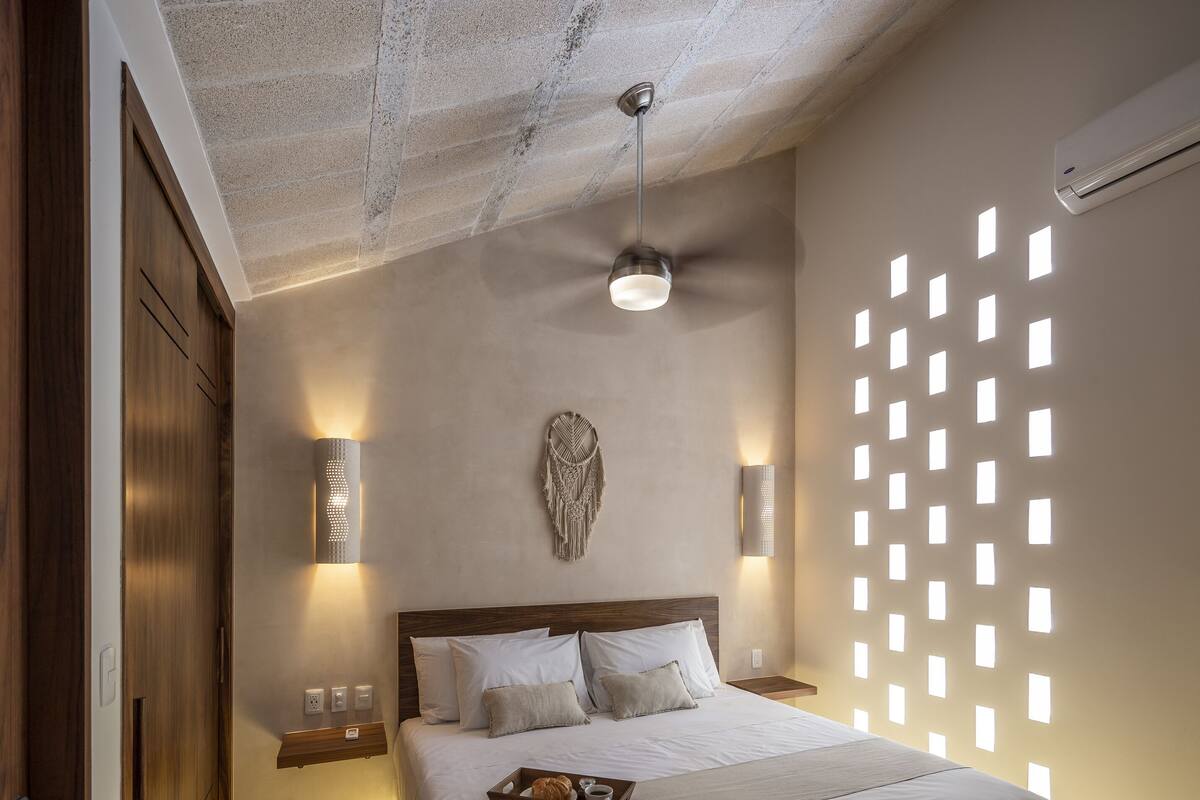 A modern bedroom with a large bed, a ceiling fan, wall-mounted lights, and a decorative wall piece.