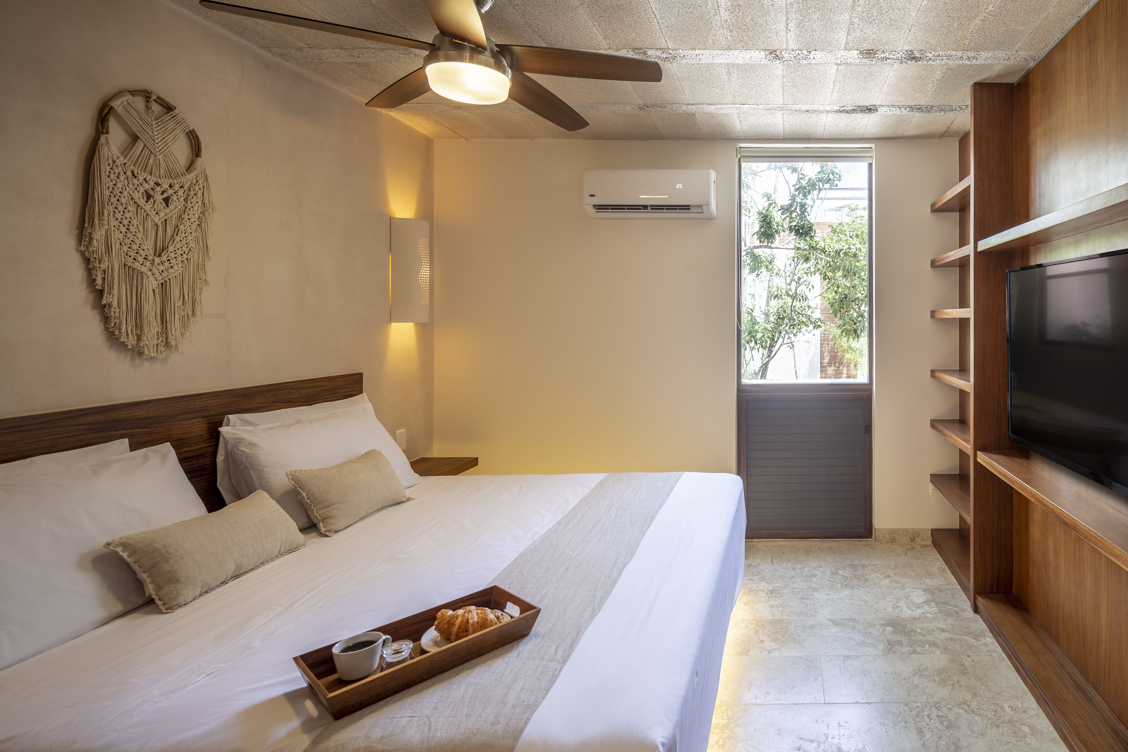 Habitación de hotel con cama, ventilador de techo, aire acondicionado de pared, televisor en un estante de madera y ventana con vista a la vegetación.