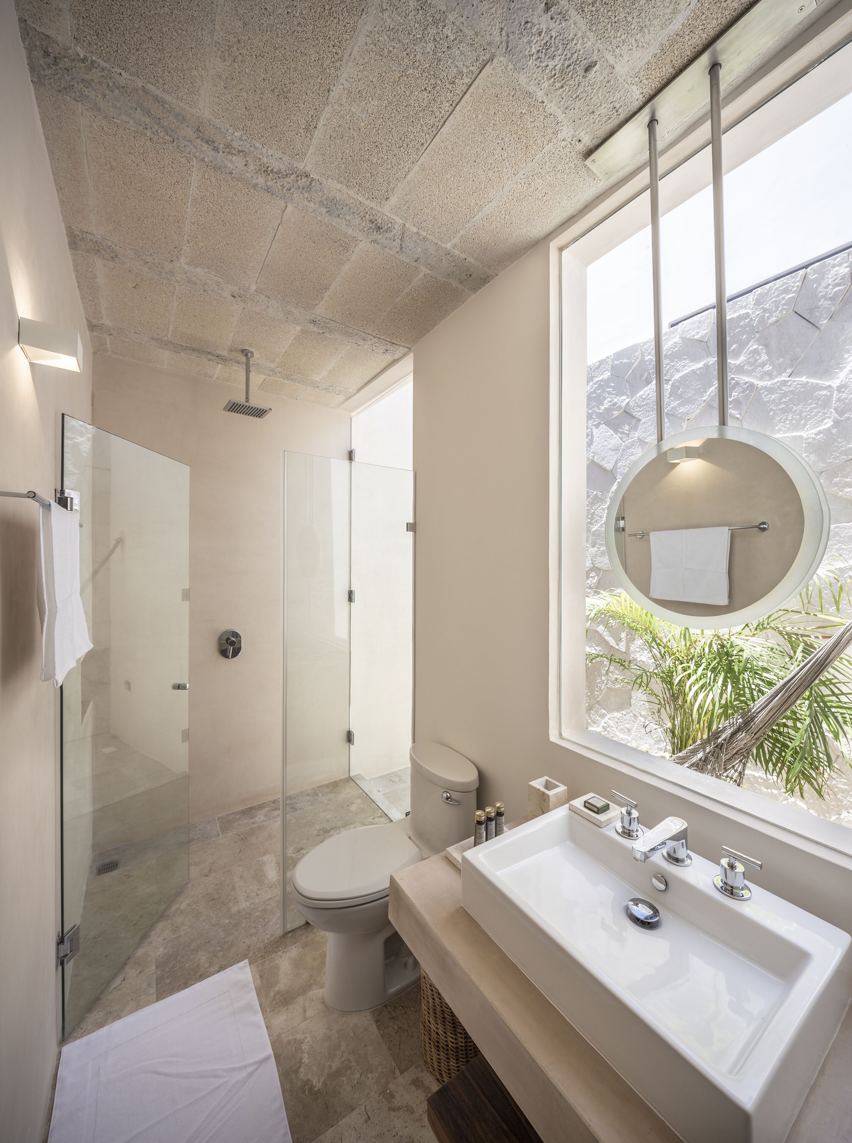 A modern bathroom with a glass-enclosed shower, a toilet, a sink with a large mirror, and a window with a view of greenery.