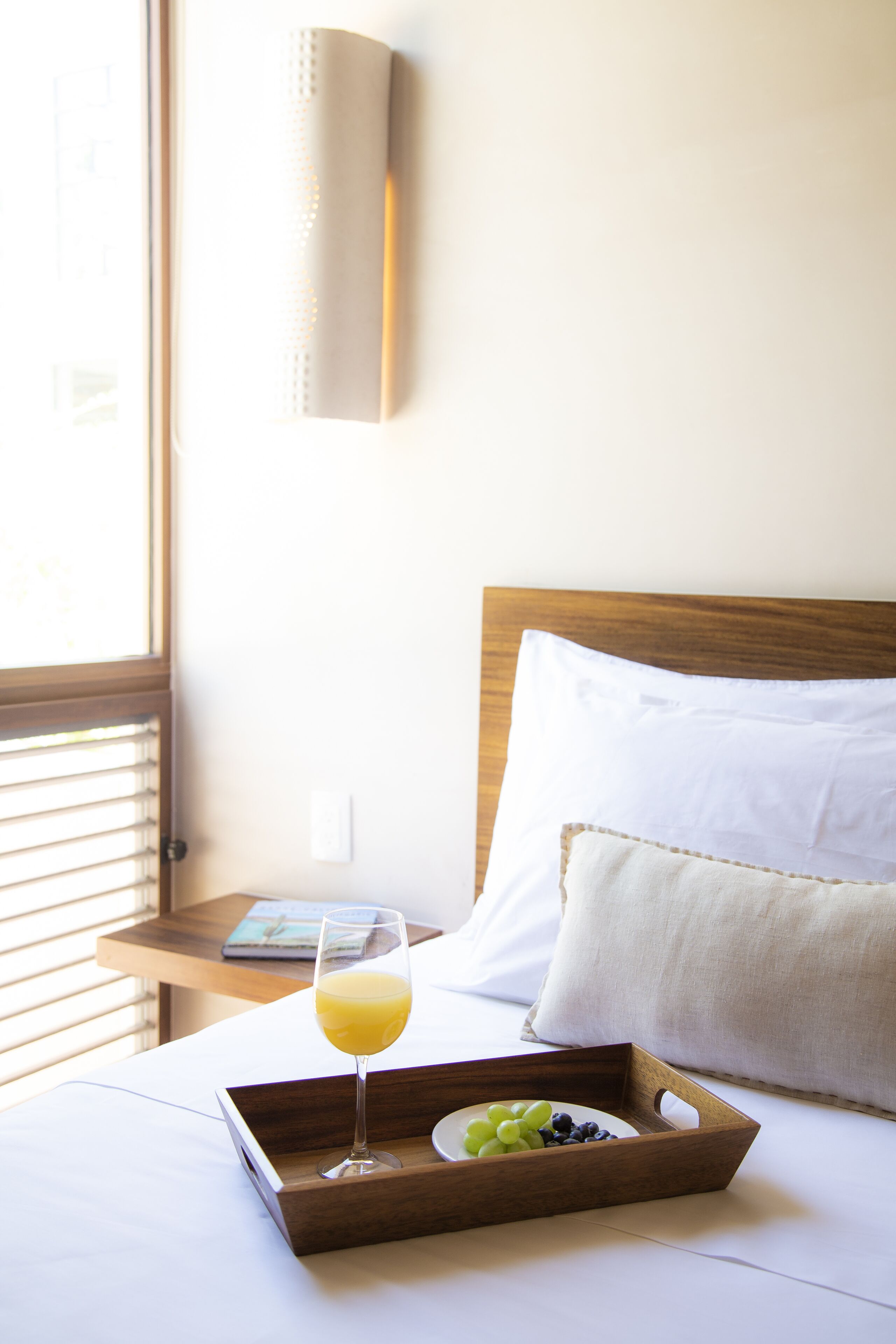 A neatly made bed with a tray containing a glass of orange juice and a bowl of fruit, a wooden nightstand, and a window with blinds.