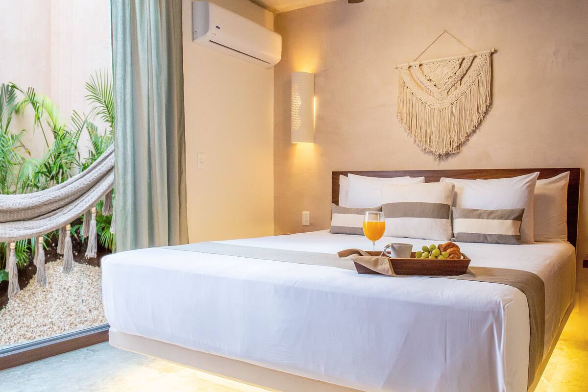 A hotel room with a bed, a bedside table with a fruit bowl and a glass of orange juice, a wall-mounted air conditioner, a macrame wall hanging, and a hammock outside the window.