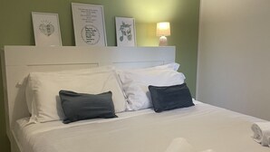 Een net opgemaakt bed met witte en donkergrijze kussens, een wit hoofdbord en een muur met ingelijste kunstwerken.