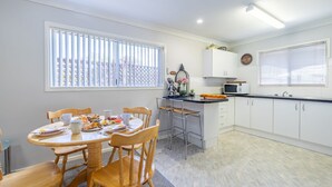 A compact kitchen with white cabinets, a microwave, and a dining area with wooden chairs and a table set for breakfast. - Lorikeet Retreat, 2/117 Tomaree Rd -  Pet Friendly, Air Conditioned Holiday House (Shoal Bay)