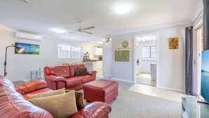 A spacious living area with a red leather sofa, a ceiling fan, and a view into the kitchen and laundry area. - Lorikeet Retreat, 2/117 Tomaree Rd -  Pet Friendly, Air Conditioned Holiday House (Shoal Bay)