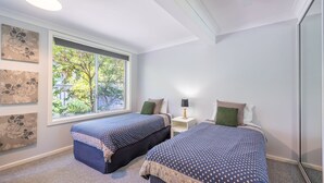A bedroom with two beds, a window with a view of greenery, and artwork on the walls. - Lorikeet Retreat, 2/117 Tomaree Rd -  Pet Friendly, Air Conditioned Holiday House (Shoal Bay)