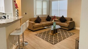 Ein modernes Wohnzimmer mit Glastisch, beiger Couch und Küchenbereich mit weißen Schränken und einem Barhocker.
