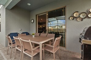 Une terrasse extérieure avec une table et des chaises en bois, un barbecue et une porte vitrée donnant sur un espace cuisine.