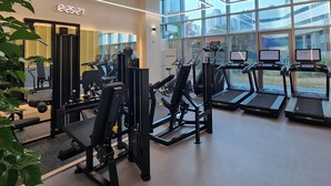 A modern gym with various exercise equipment, large windows, and a digital clock displaying 12:25:27. - Staz Hotel Premier Dongtan (Hwaseong)