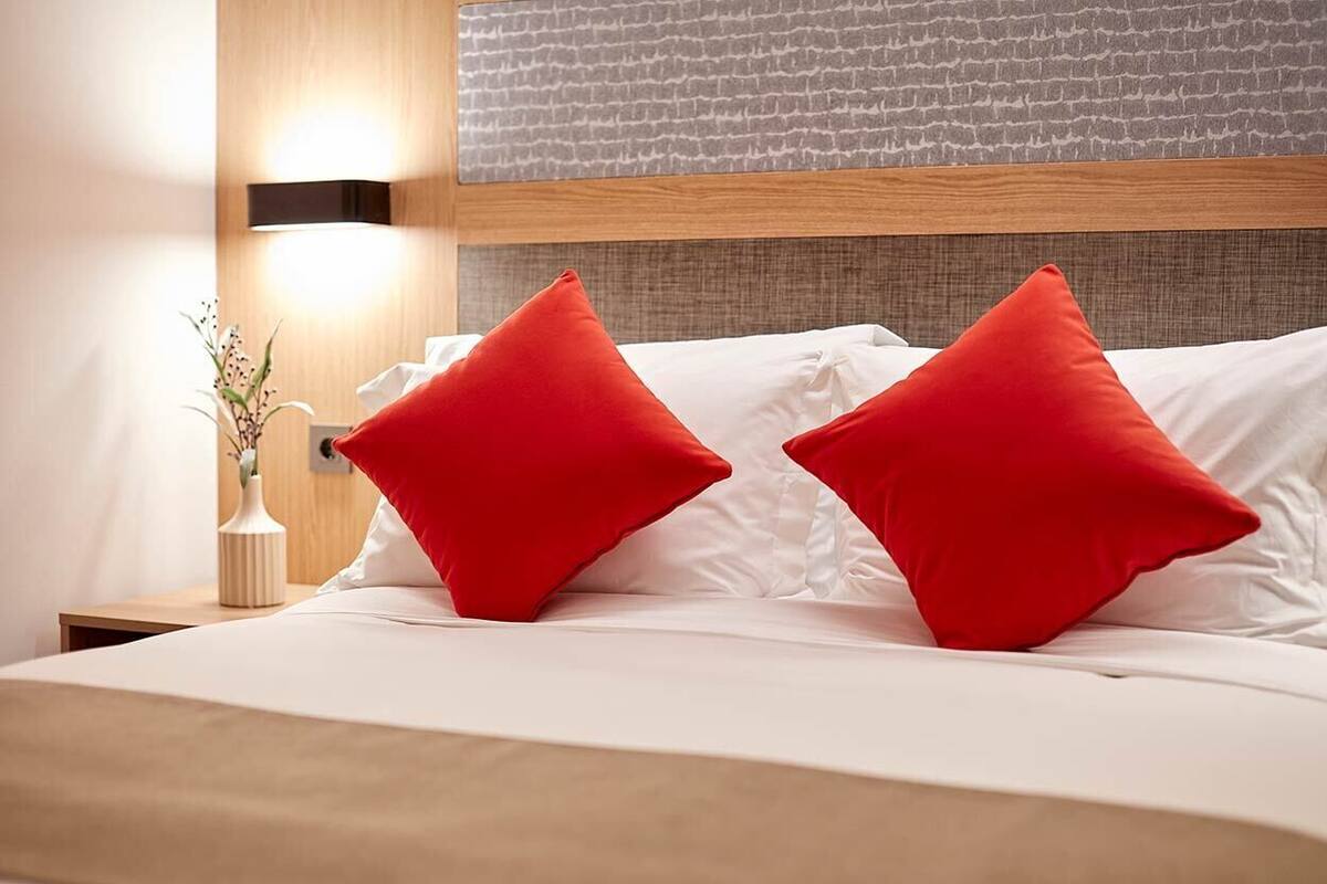 A neatly made bed with white linens and two red pillows, a bedside table with a vase of dried flowers, and a wall-mounted light fixture.
