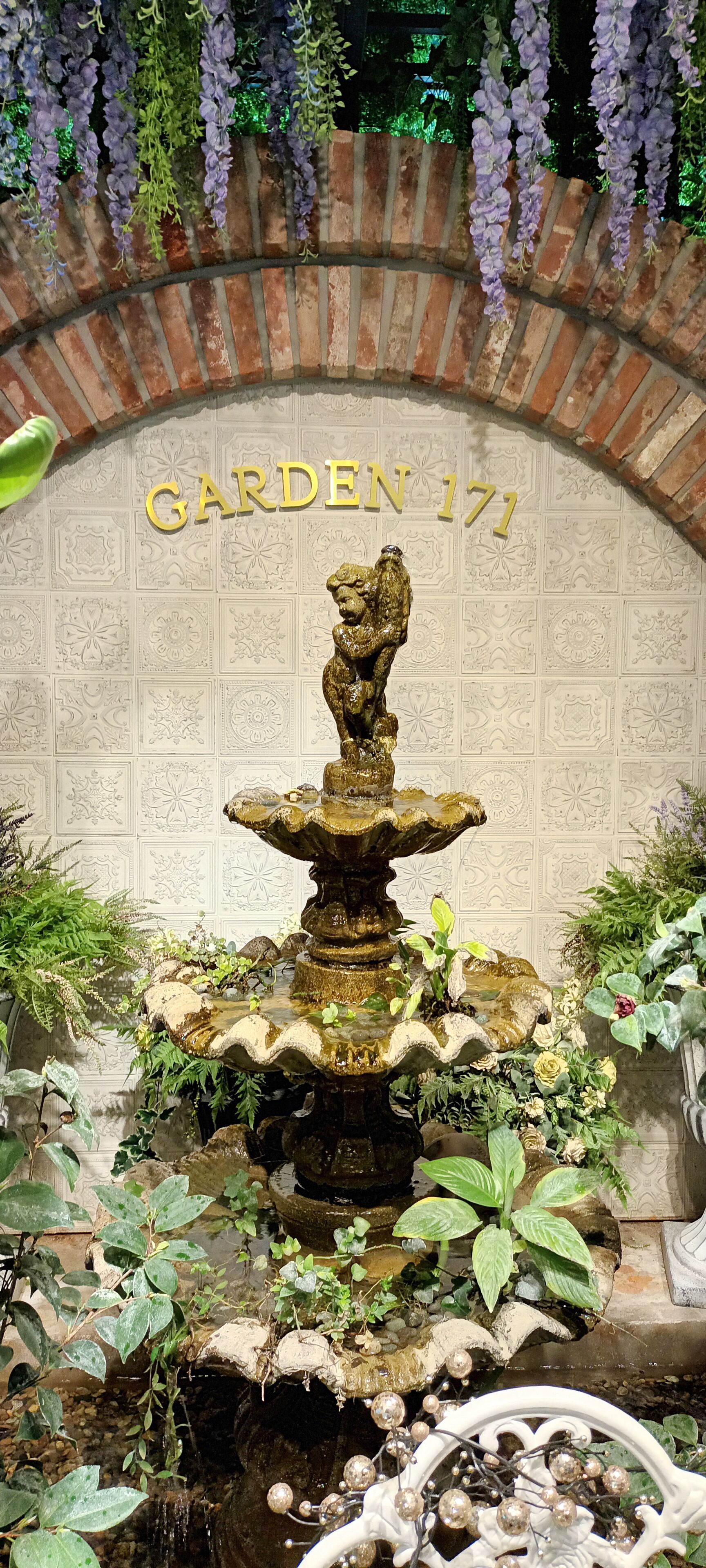Una fuente en un jardín con un arco de ladrillo con la inscripción 'GARDEN 171' y flores púrpuras colgantes.