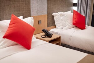 A hotel room with two beds, each with white bedding and red pillows. A bedside table with a telephone is visible.