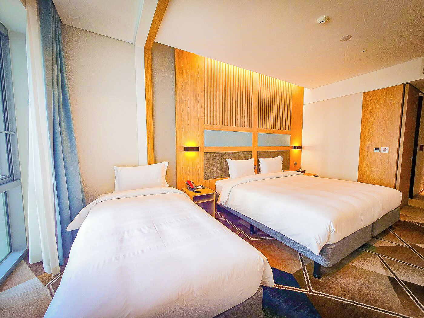 A hotel room with two beds, a wooden accent wall, a nightstand with a phone, and a window with curtains.