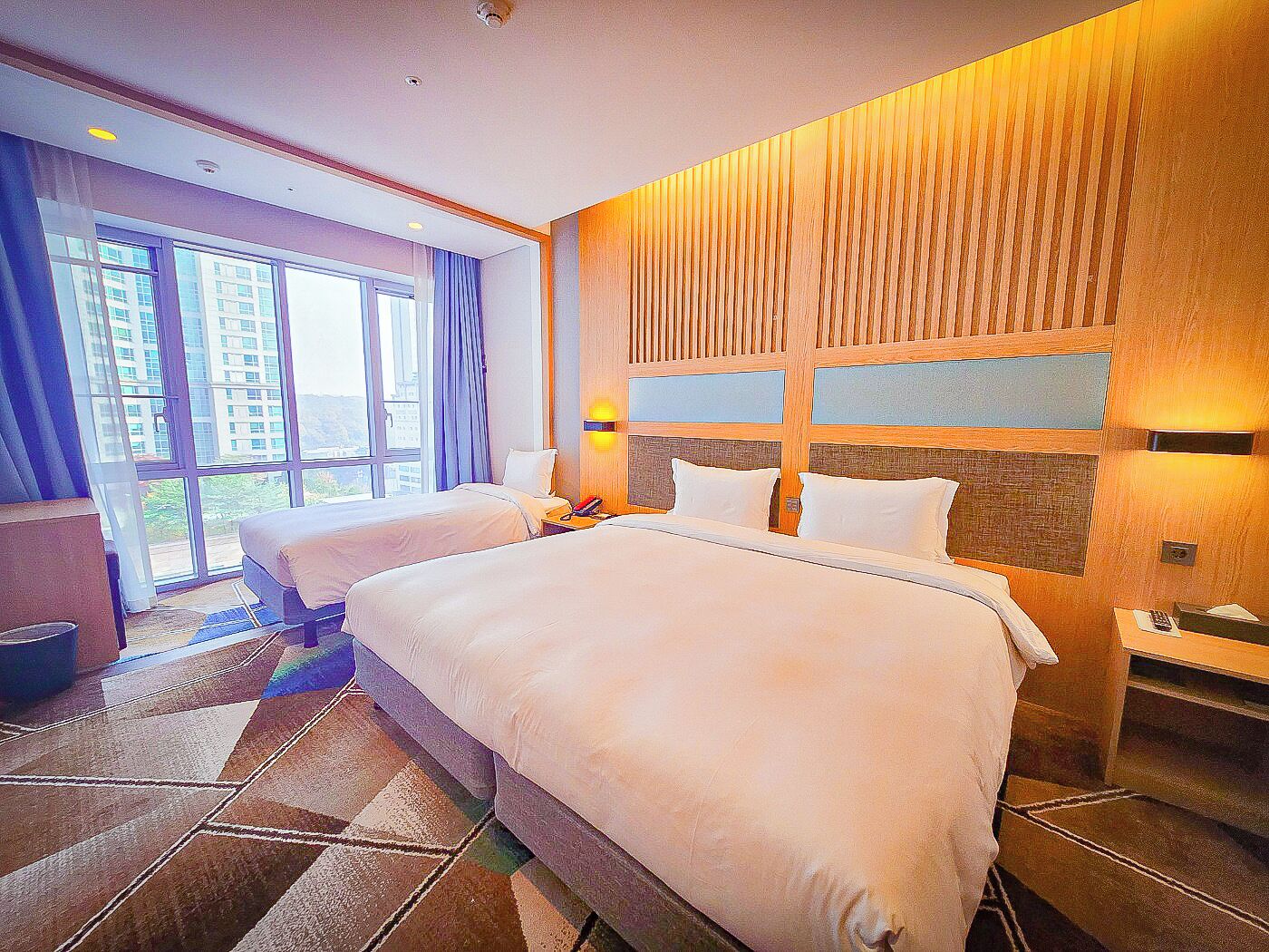 A hotel room with two beds, a large window, and a view of buildings outside.