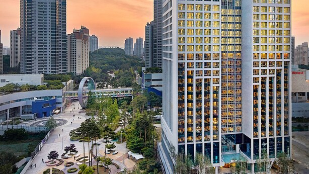 A high-rise hotel with a modern glass facade, surrounded by other tall buildings and a park area with walking paths and greenery. - Staz Hotel Premier Dongtan (Hwaseong)