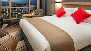 A hotel room with a large bed, red pillows, a nightstand, a chair, and a view of a cityscape through large windows. - Staz Hotel Premier Dongtan (Hwaseong)