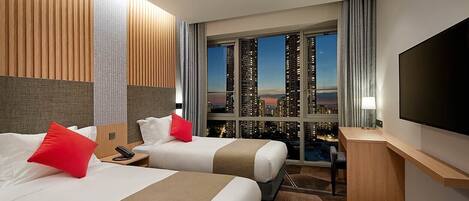 A hotel room with two beds, a TV, a desk, and a view of a cityscape at night.