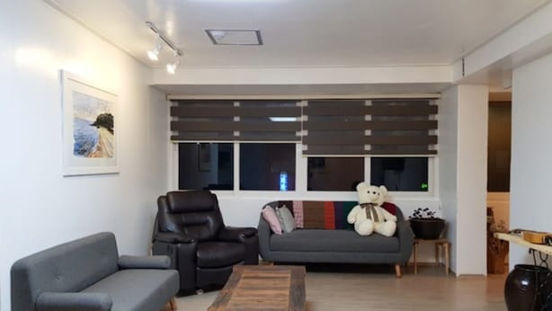 Una sala de estar con un sofá gris, un sillón de cuero negro, una mesa de centro de madera y un sofá gris. Se ve un ventanal con persianas, un peluche en el sofá y un cuadro en la pared.