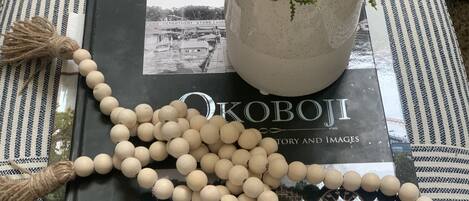 Un libro titulado 'Okoboji History and Images', una cadena de cuentas de madera y una planta en maceta sobre un cojín a rayas.