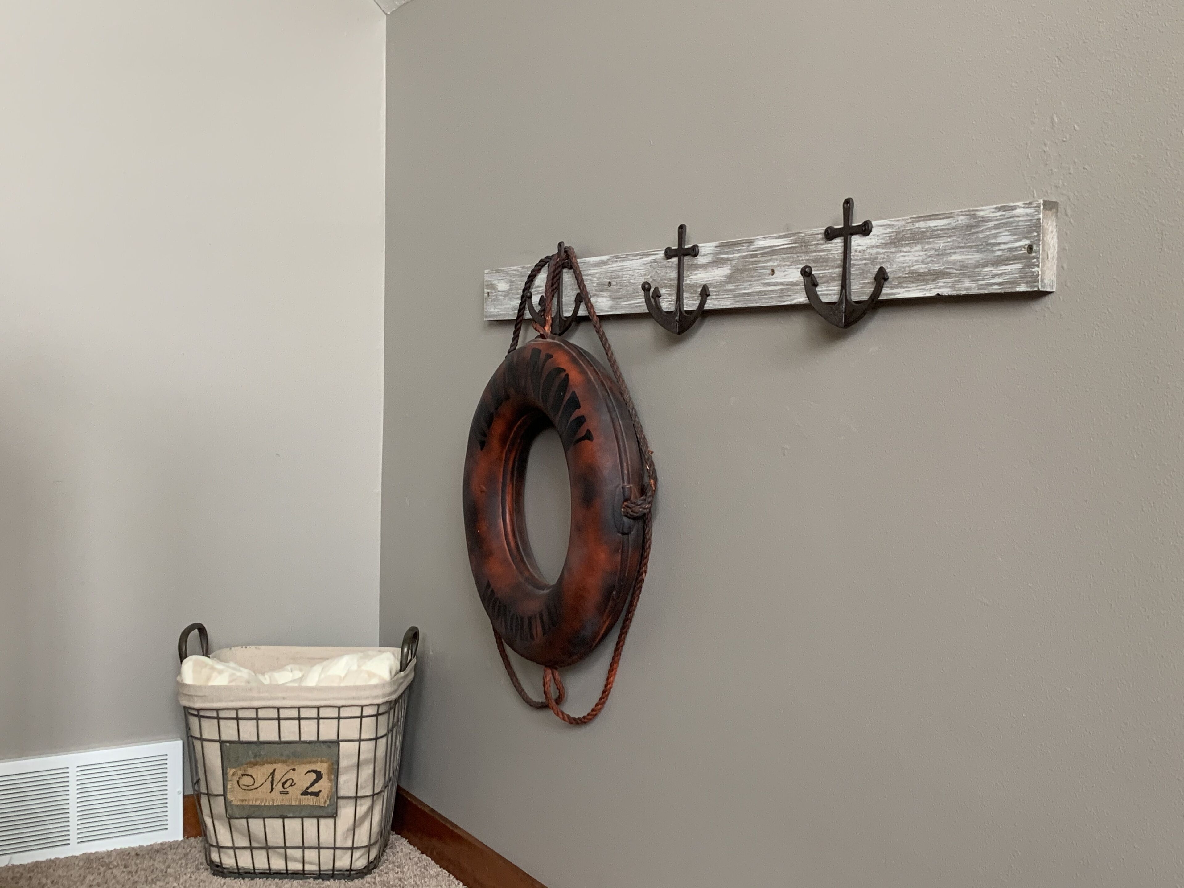 Un coin avec un support métallique fixé au mur, sur lequel sont accrochés un gilet de sauvetage et des crochets, un panier métallique avec une étiquette, et une grille d’aération.