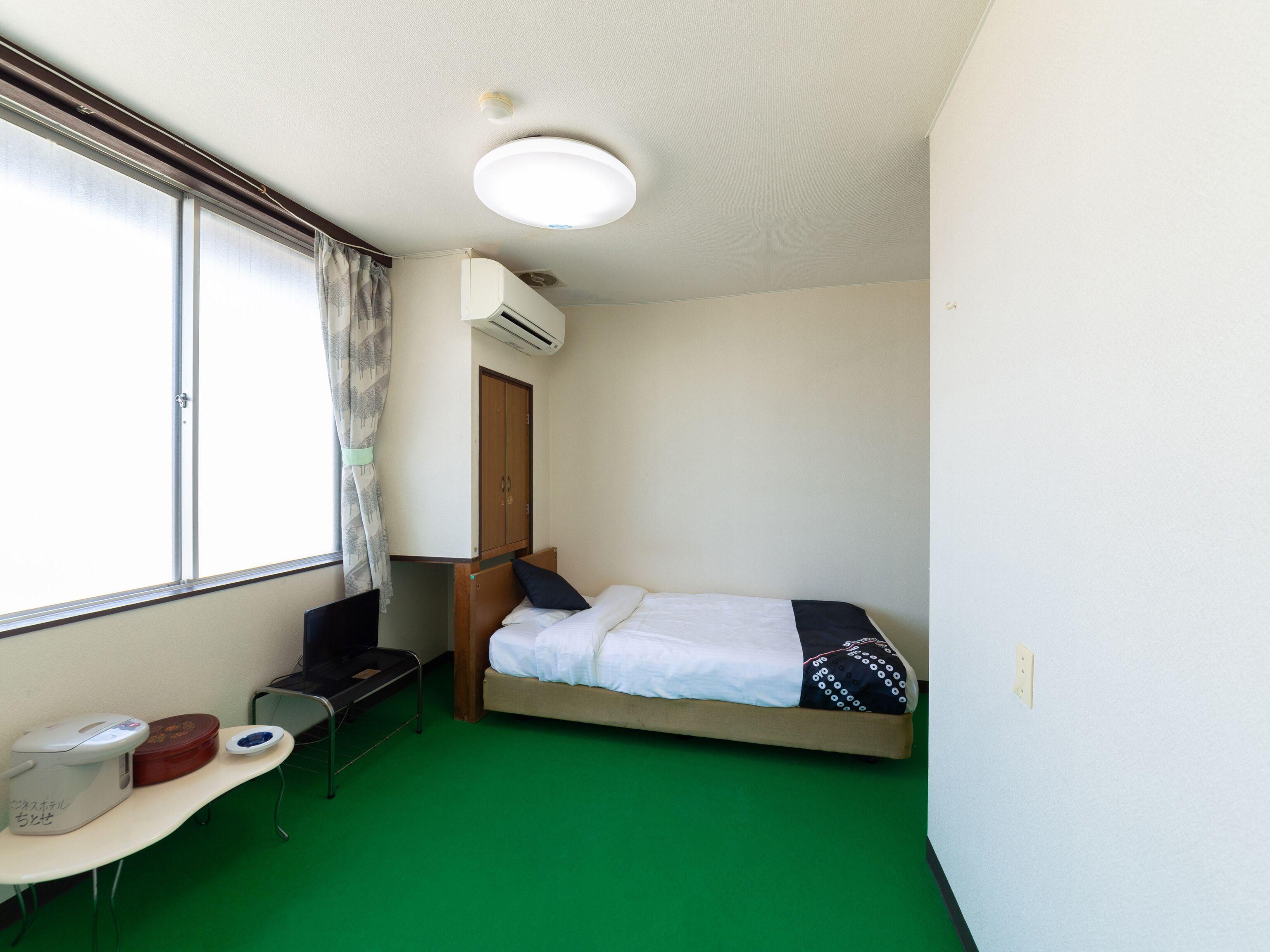 Una habitación pequeña con cama individual, alfombra verde, un escritorio con computadora y tetera, una ventana con cortinas y un equipo de aire acondicionado.