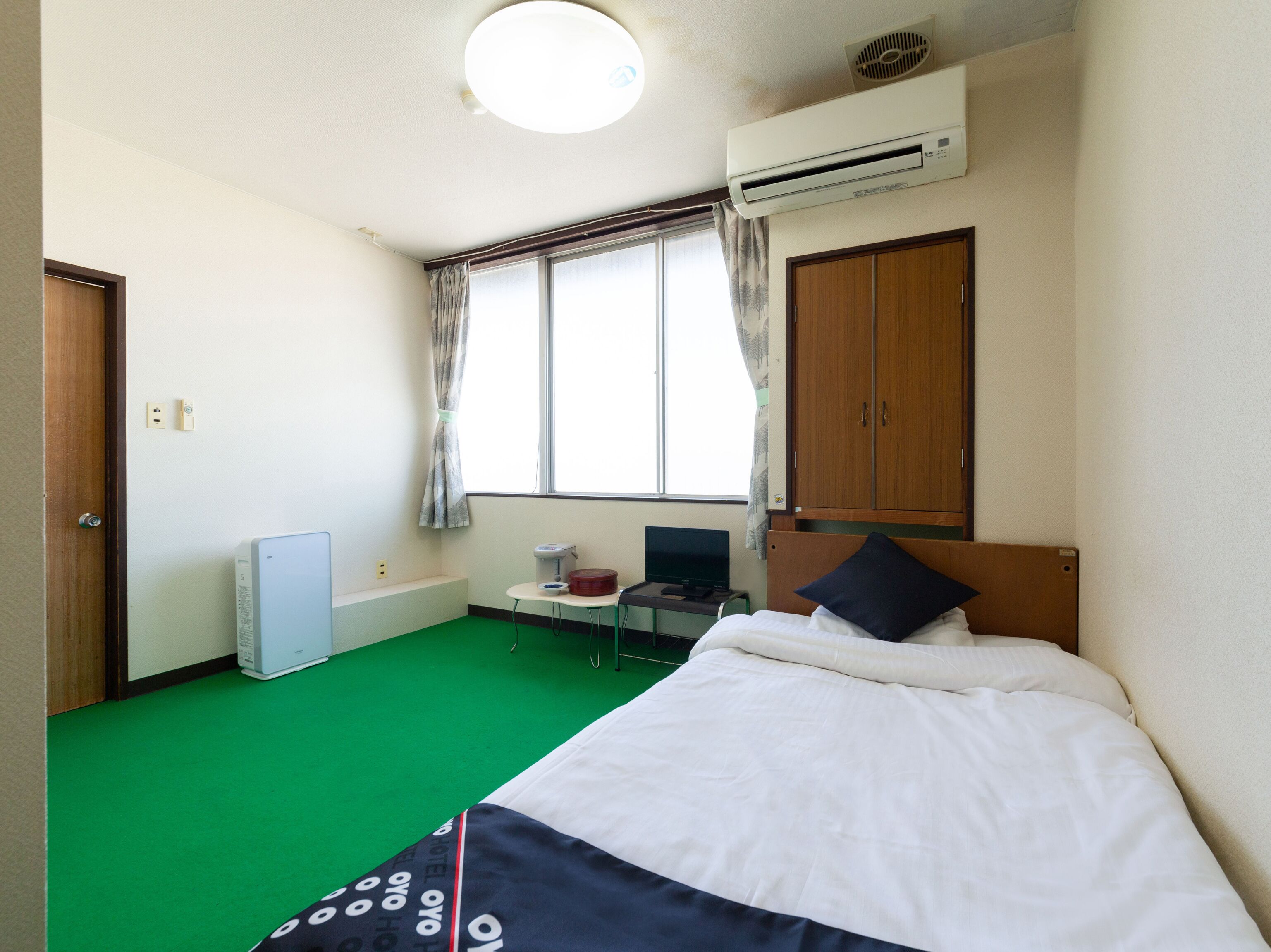 Habitación de hotel con cama, escritorio con computadora, aire acondicionado, ventana con cortinas y alfombra verde.