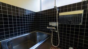 Un baño con pared de azulejos negros, una bañera de acero inoxidable, un cabezal de ducha manual y un estante pequeño con artículos de aseo.