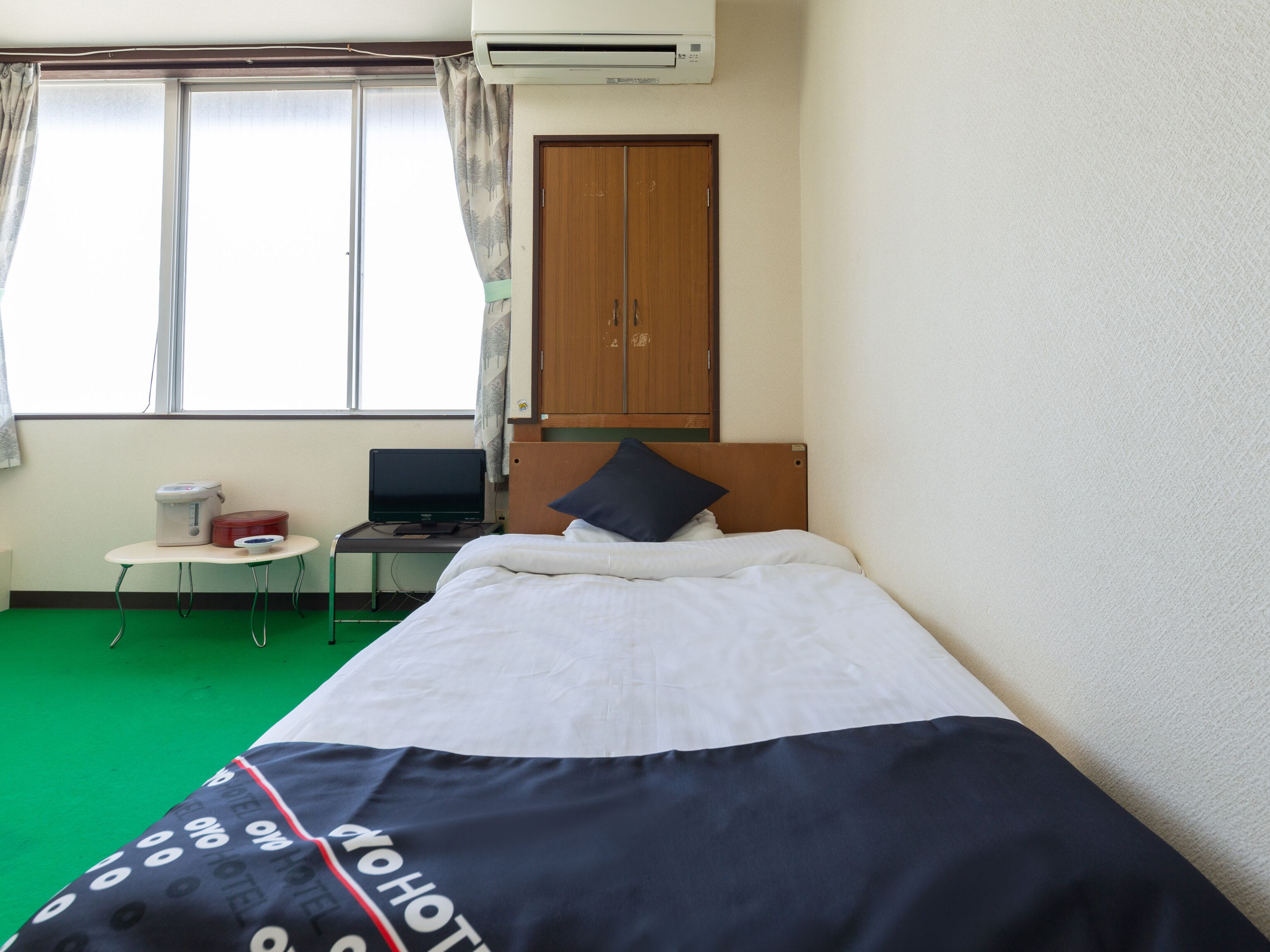 Ein Hotelzimmer mit einem Bett, einem Schreibtisch mit Computer, einem Fenster mit Vorhängen und einer Klimaanlage.